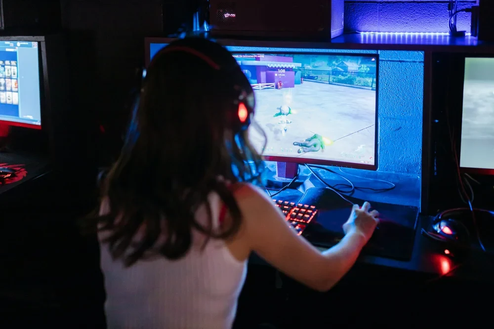 The back of a woman playing on a gaming laptop in a dark room with two screens on either side of her
