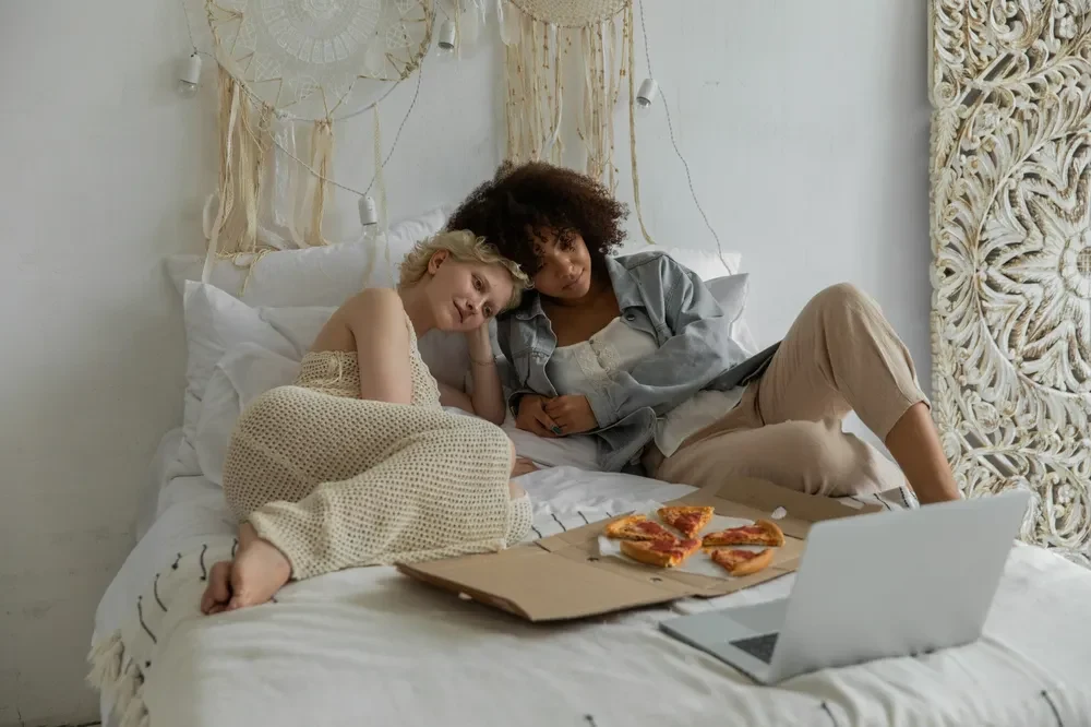 An image of two people lying on a bed, eating pizza and watching the laptop screen