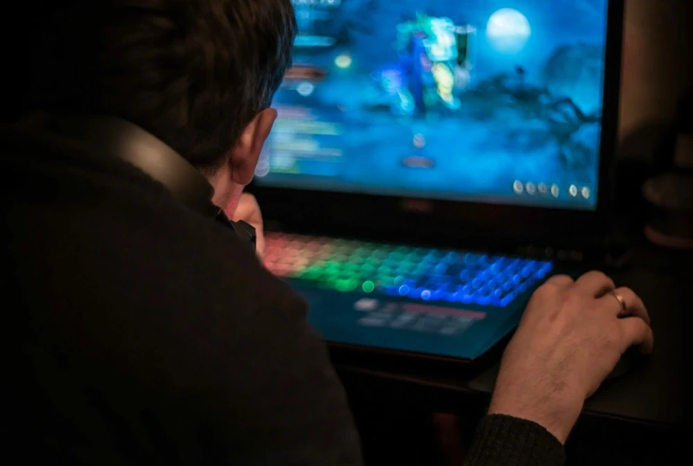 A man sits at a laptop and playing a video game in dark room. Young gamer playing video game wearing headphone.