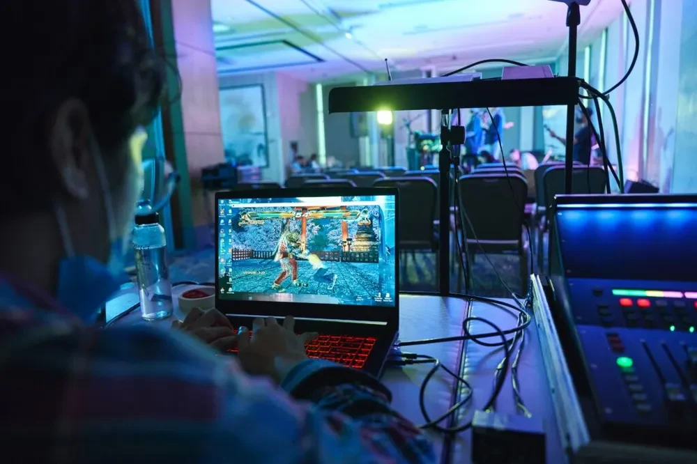 A view of the back of a person playing video games on a laptop, displaying a red keyboard colour, with a sound system on the right, chairs and other objects in the front of the image